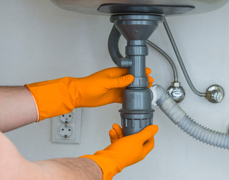 Plumber in orange rubber gloves clearing a clogged drain under the sink