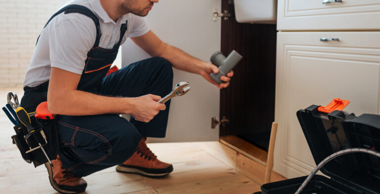 Cut view of plumber sitting in squad position in kitchen. He hold piece of pipe and wrench. Guy look under sink. Opened toolbox on floor